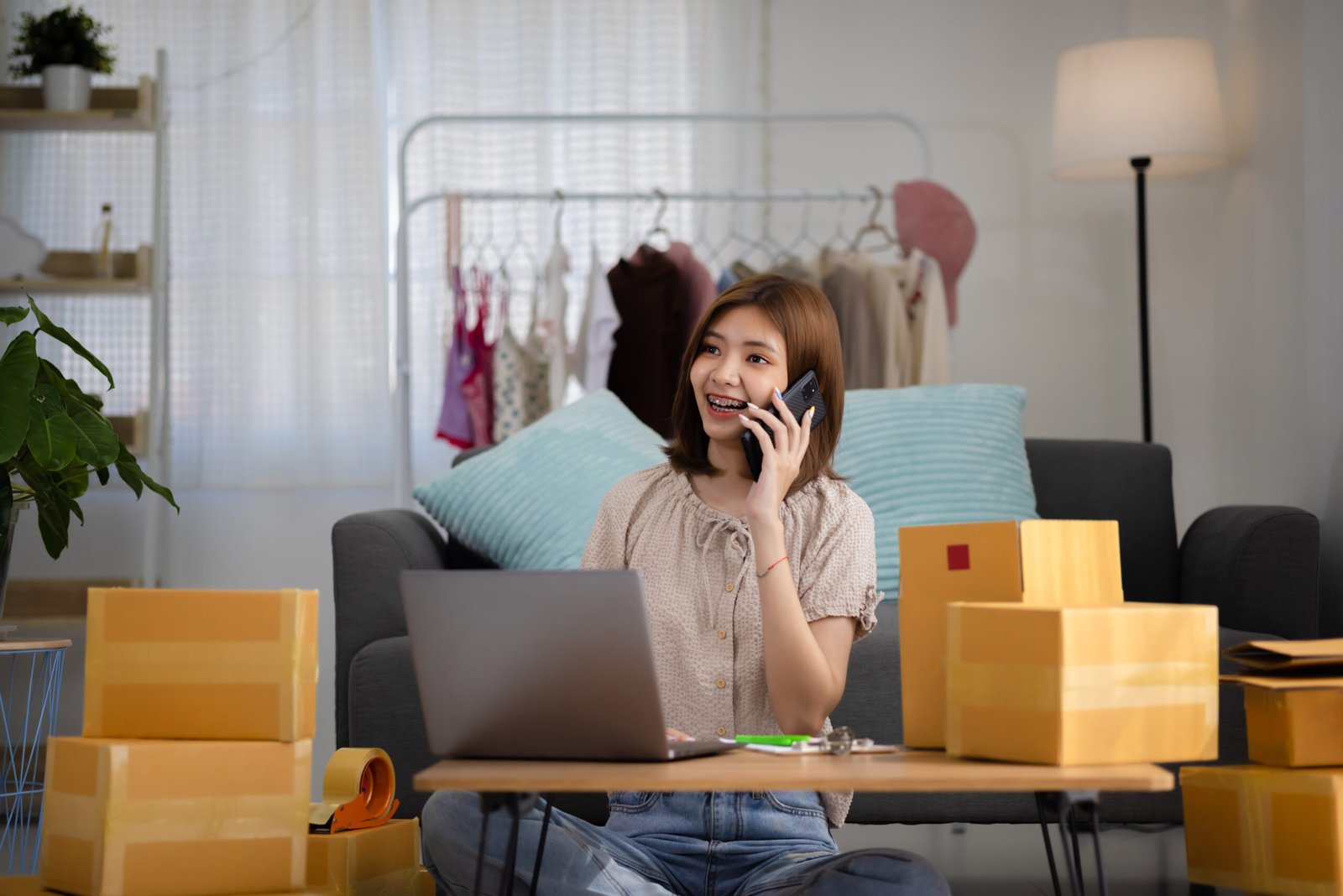 Smiling happy young Asian woman entrepreneur receiving phone call  for new sales order among boxes of product with laptop computer. Success online business and eCommerce concept.